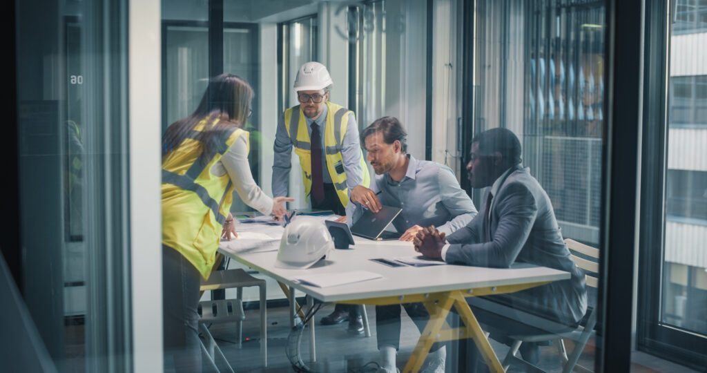 The Most Valuable Person on a Development Team Usually Isn't Who You Think executives and construction consultants meet in a glass office to review blueprints, align budgets, set milestones, and plan risk, compliance, and stakeholder communication strategies for business